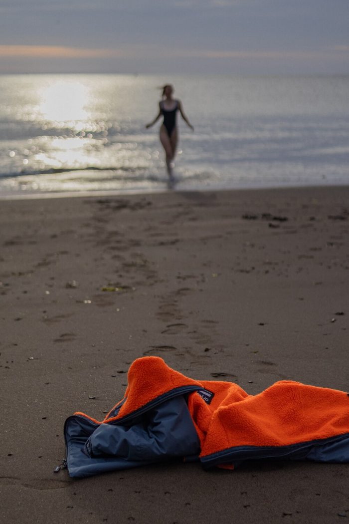 How to clean a Dryrobe. Woman running in from a swim in the sea to her orange and grey Dryrobe left on the sand.