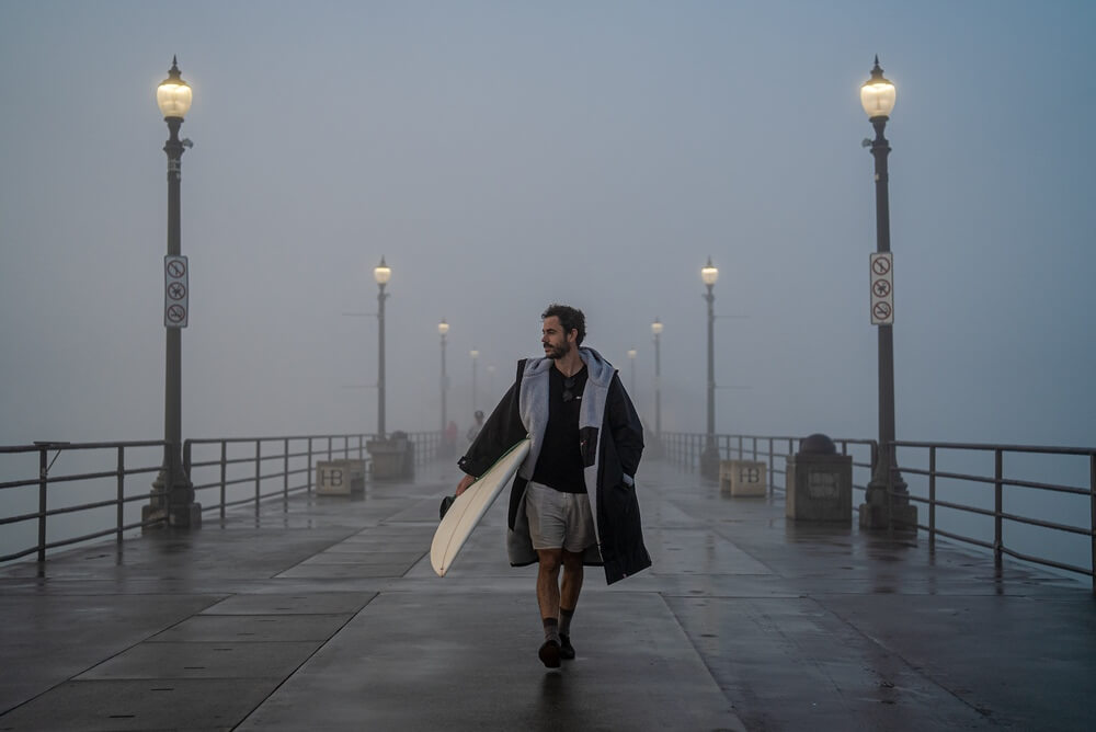 What is a Dryrobe for? Man walking down a foggy pier wearing a black and grey Dryrobe carrying a surfboard.